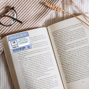 May include: An open book with text, a pair of eyeglasses, and a library card bookmark. The bookmark is light blue with the text "Library Card" and "Spooky Book Club". A sprig of wheat is placed on the book.