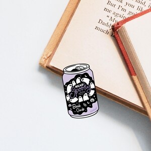 May include: A purple and white bookmark shaped like a soda can, featuring the text "Read Spooky Book Club" and ghost illustrations. The bookmark rests on open books with beige pages and red spines.