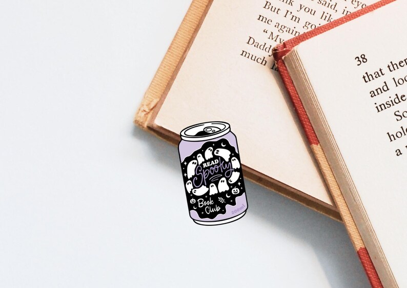 May include: A purple and white bookmark shaped like a soda can, featuring the text "Read Spooky Book Club" and ghost illustrations. The bookmark rests on open books with beige pages and red spines.