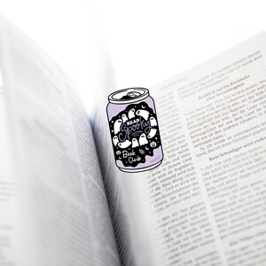 May include: A purple can-shaped sticker with the words "READ SPOOKY Book Club" and ghost illustrations. The sticker is placed on an open book with German text.