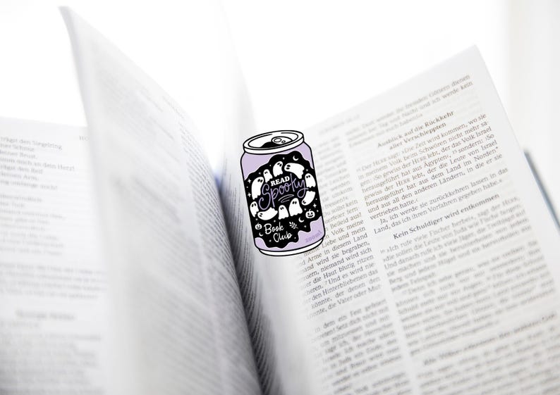 May include: A purple can-shaped sticker with the words "READ SPOOKY Book Club" and ghost illustrations. The sticker is placed on an open book with German text.