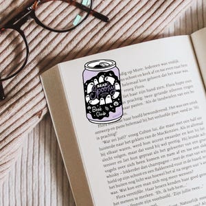 May include: A purple can-shaped sticker with the words "Read Spooky Book Club" and ghost illustrations is placed on an open book. The image includes eyeglasses and a textured surface.