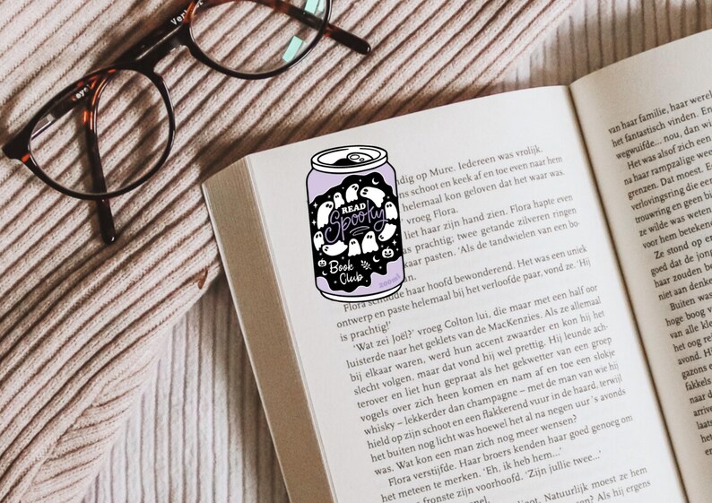 May include: A purple can-shaped sticker with the words "Read Spooky Book Club" and ghost illustrations is placed on an open book. The image includes eyeglasses and a textured surface.