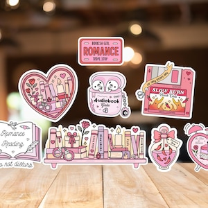 May include: A collection of pink and white stickers with a romance theme. Designs include books, hearts, and the text "Romance Reading", "Bookish Girl Romance Trope Stop", and "Slow Burn Book Club".