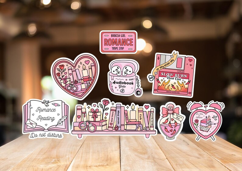 May include: A collection of pink and white stickers with a romance theme. Designs include books, hearts, and the text "Romance Reading", "Bookish Girl Romance Trope Stop", and "Slow Burn Book Club".