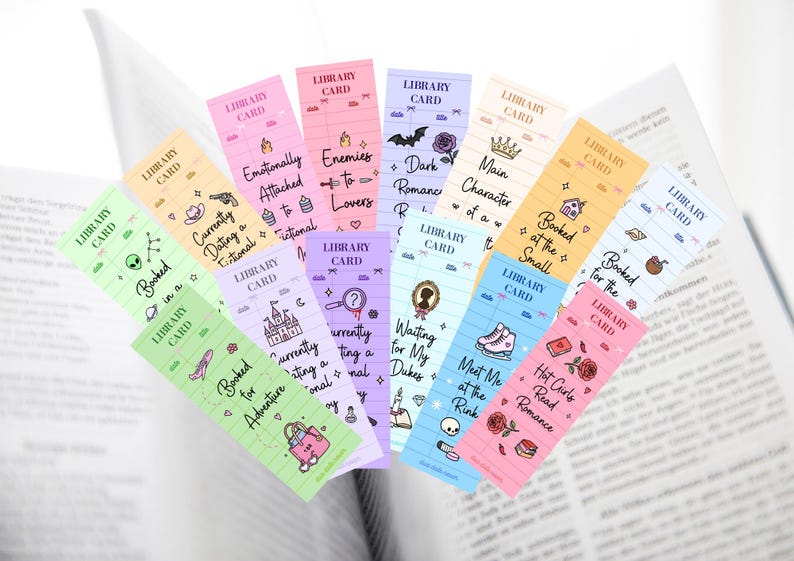 May include: A collection of colorful bookmarks with the words "Library Card" at the top. Each bookmark features a different theme, including "Booked in a Galaxy," "Enemies to Lovers," and "Hot Girls Read Romance."