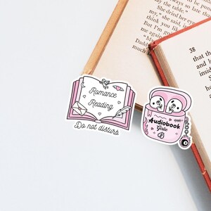 May include: Pink stickers with the text "Romance Reading Do not disturb" and "Audiobook Girlie". The stickers are placed on open books. The audiobook sticker shows a pair of earbuds with flowers.