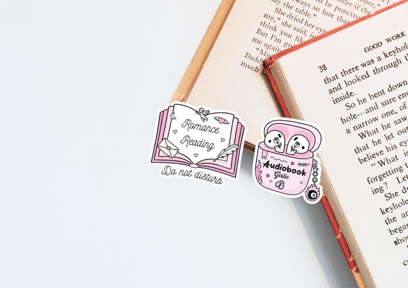 May include: Pink stickers with the text "Romance Reading Do not disturb" and "Audiobook Girlie". The stickers are placed on open books. The audiobook sticker shows a pair of earbuds with flowers.