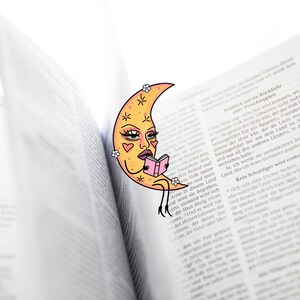 May include: A whimsical bookmark featuring a crescent moon with a face, reading a small pink book. The bookmark is placed in an open book with text. The moon has a yellow face with pink hearts and flower accents.