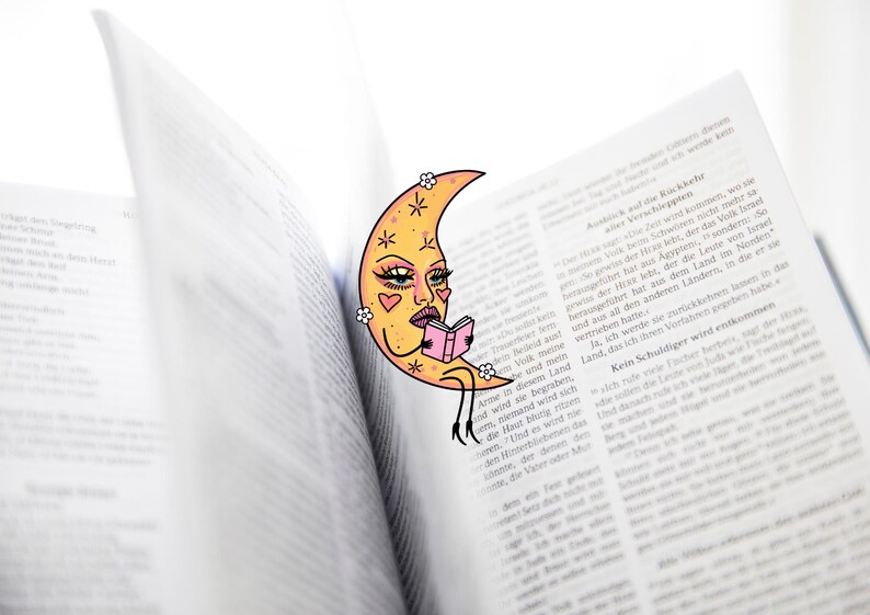 May include: A whimsical bookmark featuring a crescent moon with a face, reading a small pink book. The bookmark is placed in an open book with text. The moon has a yellow face with pink hearts and flower accents.