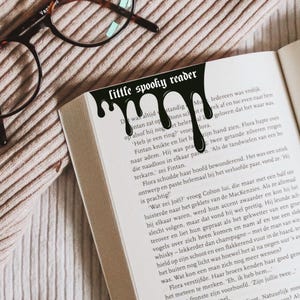 Pode incluir: Um marcador de livro preto com o texto "little spooky reader" e um design gotejante, colocado em um livro aberto. O livro est&aacute; em uma superf&iacute;cie texturizada, marrom claro, com &oacute;culos no fundo.
