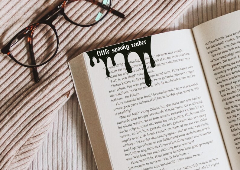 Pode incluir: Um marcador de livro preto com o texto "little spooky reader" e um design gotejante, colocado em um livro aberto. O livro est&aacute; em uma superf&iacute;cie texturizada, marrom claro, com &oacute;culos no fundo.