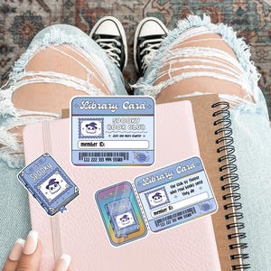 May include: A collection of stickers and a notebook. The stickers include a blue library card with the text "Spooky Book Club" and a cartoon ghost. A pink notebook and a spiral-bound notebook are also visible.