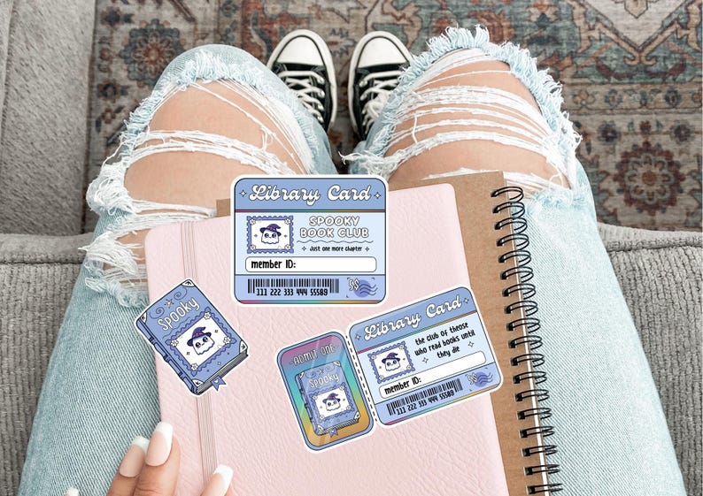 May include: A collection of stickers and a notebook. The stickers include a blue library card with the text "Spooky Book Club" and a cartoon ghost. A pink notebook and a spiral-bound notebook are also visible.