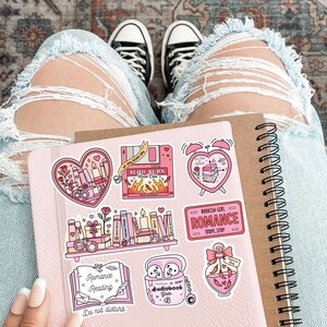 May include: A pink notebook with spiral binding and several stickers featuring romantic themes. The stickers include hearts, books, candles, and the text "Romance Reading". The background includes distressed blue jeans and black and white sneakers.