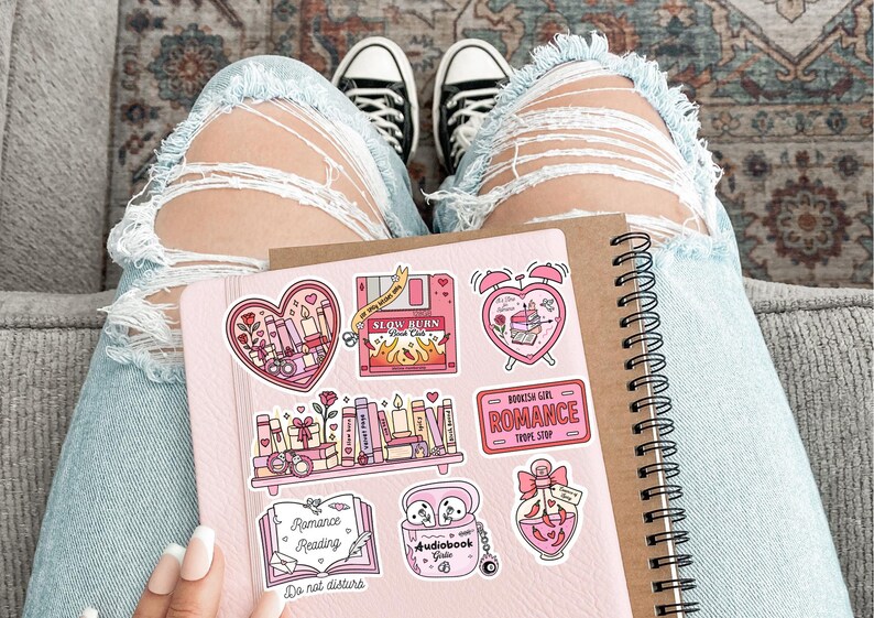 May include: A pink notebook with spiral binding and several stickers featuring romantic themes. The stickers include hearts, books, candles, and the text "Romance Reading". The background includes distressed blue jeans and black and white sneakers.