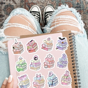 May include: A spiral-bound notebook with a light pink cover featuring eleven individual stickers. Each sticker depicts a stack of books with the text 'It's Time' and a unique icon on top, including a candle, skull, house, bat, UFO, magic 8-ball, teapot, dragon, plane, crab, and roller skates.