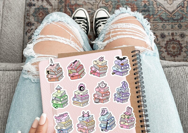 May include: A spiral-bound notebook with a light pink cover featuring eleven individual stickers. Each sticker depicts a stack of books with the text 'It's Time' and a unique icon on top, including a candle, skull, house, bat, UFO, magic 8-ball, teapot, dragon, plane, crab, and roller skates.