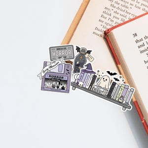 May include: A selection of Halloween-themed stickers. The stickers include a bookshelf, a tombstone, a floppy disk, and a sign that says "HORROR". The colour palette is purple, grey, and black, with a ghost and bats.