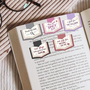 Puede incluir: P&aacute;gina de libro con varios marcadores decorativos. Cada marcador tiene la forma de un libro abierto y presenta textos como "trust NO ONE" y "this got SPICY fast". Los marcadores son de varios colores, incluyendo rosa, morado y rojo.
