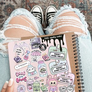 Pode incluir: Um caderno espiral com uma capa rosa com v&aacute;rios adesivos com um tema assustador. Os adesivos incluem frases como "Read Books" e "Probably Reading Thriller". O caderno &eacute; segurado no colo de uma pessoa, com jeans rasgados azul claro e t&ecirc;nis preto e branco.
