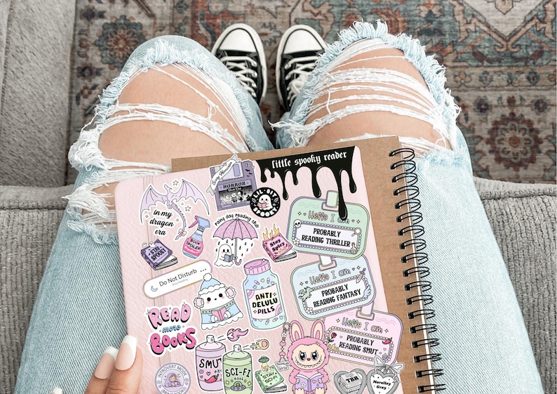 Pode incluir: Um caderno espiral com uma capa rosa com v&aacute;rios adesivos com um tema assustador. Os adesivos incluem frases como "Read Books" e "Probably Reading Thriller". O caderno &eacute; segurado no colo de uma pessoa, com jeans rasgados azul claro e t&ecirc;nis preto e branco.