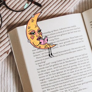 May include: A whimsical bookmark featuring a crescent moon with a face, reading a book. The bookmark is yellow with pink accents and is placed in an open book. The image also includes eyeglasses.
