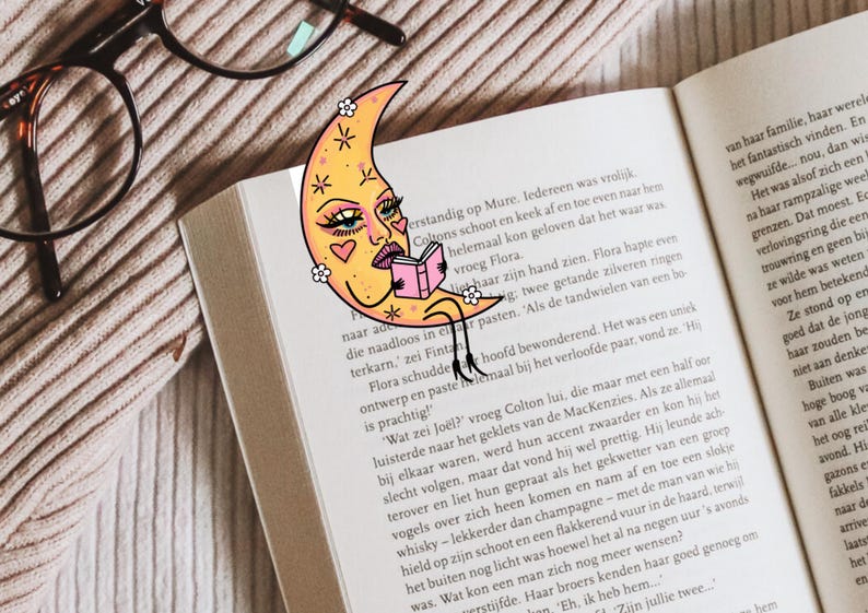 May include: A whimsical bookmark featuring a crescent moon with a face, reading a book. The bookmark is yellow with pink accents and is placed in an open book. The image also includes eyeglasses.