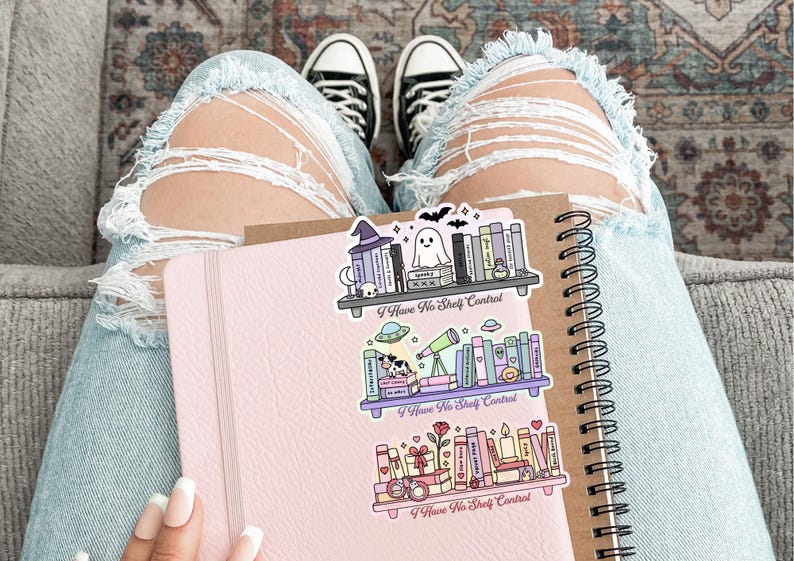 Op de afbeelding: Een roze notitieboek en een spiraalgebonden notitieboek met drie decoratieve stickers met boekenplanken met de tekst "I Have No Shelf Control". De stickers hebben een Halloween-, ruimte- en geschenkthema.