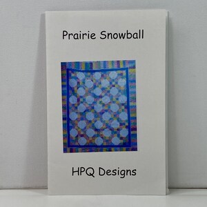 May include: A white paper pattern booklet titled Prairie Snowball by HPQ Designs. The cover features a central image of a quilt with light blue octagonal shapes surrounded by a multi-colored grid border and a solid blue frame.