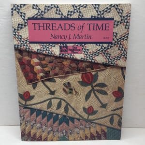 Könnte beinhalten: Ein Buch mit dem Titel "Threads of Time" von Nancy J. Martin. Das Cover zeigt ein Patchwork-Quilt-Design mit verschiedenen Mustern, darunter geometrische Formen, florale Motive und Pfeile. Die Farbpalette umfasst Beige, Rosa, Rot und Blau.