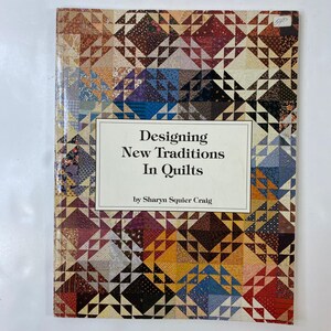 Puede incluir: Un libro titulado "Designing New Traditions In Quilts" de Sharyn Squier Craig. La portada presenta un diseño de colcha de patchwork colorido en tonos marrón, azul, rojo y crema. El libro está sobre una superficie blanca.