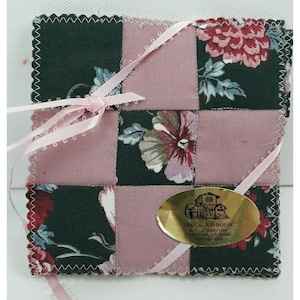 Puede incluir: Un juego de posavasos de patchwork con un estampado floral en verde oscuro, rosa y rojo. El juego está atado con una cinta rosa e incluye una etiqueta ovalada dorada con texto.