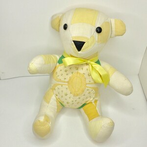 Pode incluir: Um urso de pelúcia feito à mão com um design de patchwork em tons de amarelo e creme. O urso tem olhos de botão pretos, um nariz preto e uma fita amarela em volta do pescoço. O corpo do urso apresenta um padrão floral.