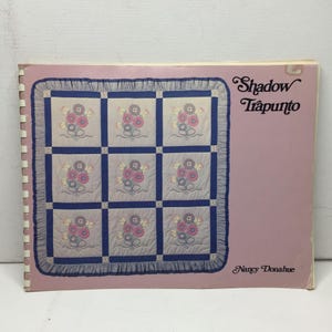 Puede incluir: Un libro encuadernado en espiral con una cubierta rosa que presenta un diseño de edredón. El patrón del edredón incluye nueve cuadrados con bordados florales en rosa, morado y amarillo. El título "Shadow Trapunto" está escrito en cursiva, y el nombre de la autora, Nancy Donahue, también es visible.