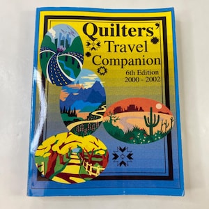 Puede incluir: Un libro titulado "Quilters' Travel Companion" con una cubierta azul y un borde amarillo. La portada presenta ilustraciones coloridas de paisajes, incluyendo carreteras, montañas y escenas desérticas. También es visible el texto "6th Edition 2000-2002".