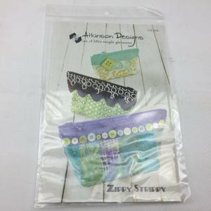 Könnte beinhalten: Eine transparente Plastikverpackung mit einem Schnittmuster für drei Stoffbeutel. Die Beutel sind in Grün-, Blau- und Lilatönen gehalten und mit dekorativen Knöpfen und gemustertem Stoff versehen. Der Text "Atkinson Designs" und "Zippy Strippy" sind sichtbar.