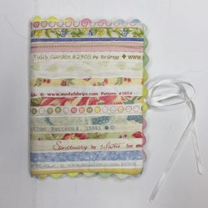 May include: A fabric-covered notebook with a scalloped edge and a white ribbon tie. The cover is made of various fabric strips with floral and patterned designs. Text includes "Tulip Garden," "Pattern #3854," and "Sanctuary."
