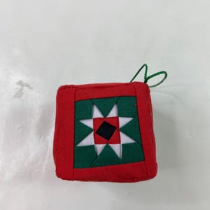 May include: A square, handmade Christmas ornament with a patchwork design. The ornament features a red border, green and white star pattern, and a black square in the center. A green string is attached for hanging.