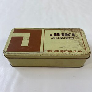 Könnte beinhalten: Eine rechteckige, cremefarbene Aufbewahrungsdose aus Metall mit einem braunen geometrischen Logo auf der linken Seite. Der Deckel trägt die Aufschrift JUKI ACCESSORIES und TOKYO JUKI INDUSTRIAL CO., LTD. in dunkelbrauner Schrift mit Gebrauchsspuren.