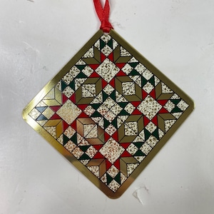 Puede incluir: Adorno navideño cuadrado con borde dorado y cinta roja para colgar. El adorno presenta un diseño tipo patchwork con formas geométricas rojas, verdes, doradas y blancas. El diseño es simétrico y festivo.