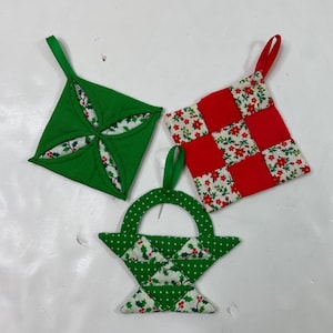 Peut inclure: Trois décorations de Noël faites à la main. L'une est un carré vert avec un motif floral, une autre est un carré patchwork rouge et blanc, et la troisième est une forme de panier vert avec une poignée. Chacune a une boucle de ruban pour accrocher.