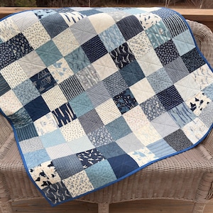 May include: A patchwork quilt with a blue border, composed of various blue and white patterned squares. The quilt is draped over a wicker chair on a wooden deck. The patterns include geometric shapes and nature-inspired designs.