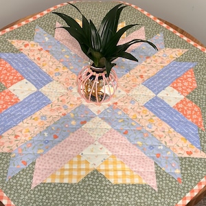 Pu&ograve; includere: Runner da tavolo a fantasia floreale con un design a stella nei toni del rosa, blu e pesca. Un piccolo vaso di filo rosa contiene una pianta verde. Il runner &egrave; su un tavolo di legno.