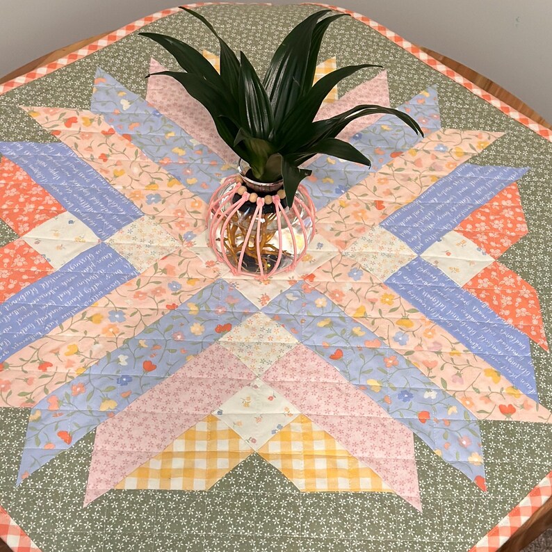 Pu&ograve; includere: Runner da tavolo a fantasia floreale con un design a stella nei toni del rosa, blu e pesca. Un piccolo vaso di filo rosa contiene una pianta verde. Il runner &egrave; su un tavolo di legno.