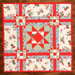 May include: A square Christmas-themed quilt with a red border. The quilt features a central red star design, surrounded by squares of fabric with festive patterns, including gift boxes, reindeer, and squirrels. The quilt is approximately 24 inches square.