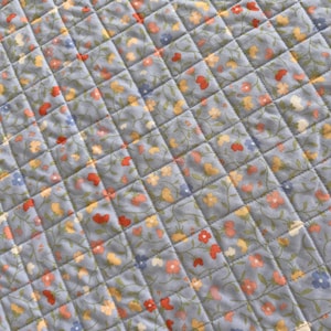Pu&ograve; includere: Primo piano di un tessuto trapuntato con motivo floreale. Il tessuto &egrave; di colore azzurro con un motivo a rombi creato dalle cuciture. Piccoli fiori nei toni del rosso, giallo e blu sono sparsi sulla superficie.