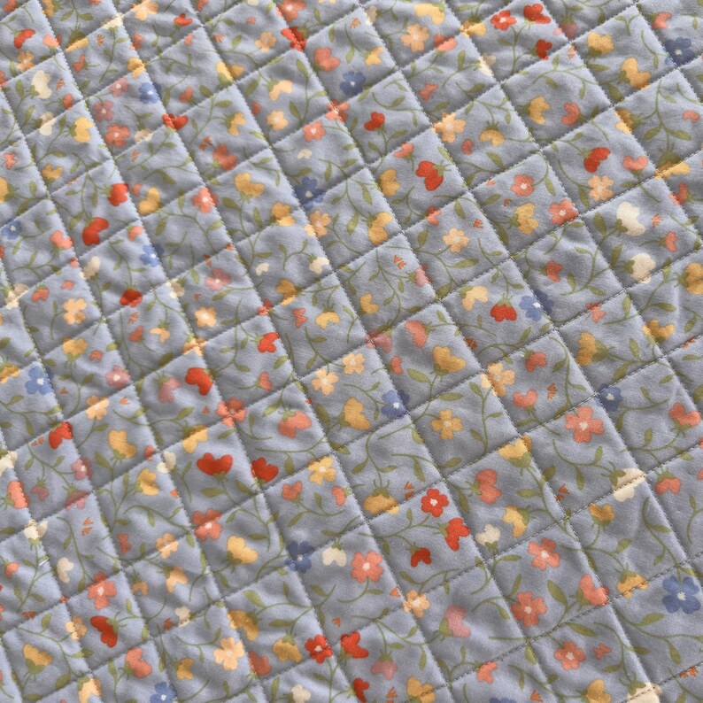 Pu&ograve; includere: Primo piano di un tessuto trapuntato con motivo floreale. Il tessuto &egrave; di colore azzurro con un motivo a rombi creato dalle cuciture. Piccoli fiori nei toni del rosso, giallo e blu sono sparsi sulla superficie.