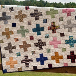 May include: A handmade quilt with a cream-colored background and a repeating pattern of colorful plus signs. The plus signs are in various colors, including blue, pink, green, and brown. The quilt has a dark blue border.