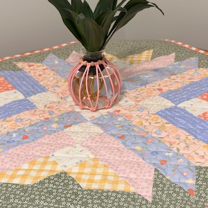 Pu&ograve; includere: Un vaso di vetro trasparente con una gabbia decorativa rosa contiene piante verdi. Il vaso &egrave; posto su una colorata trapunta patchwork con motivi floreali e geometrici nei toni del rosa, blu, giallo e verde.